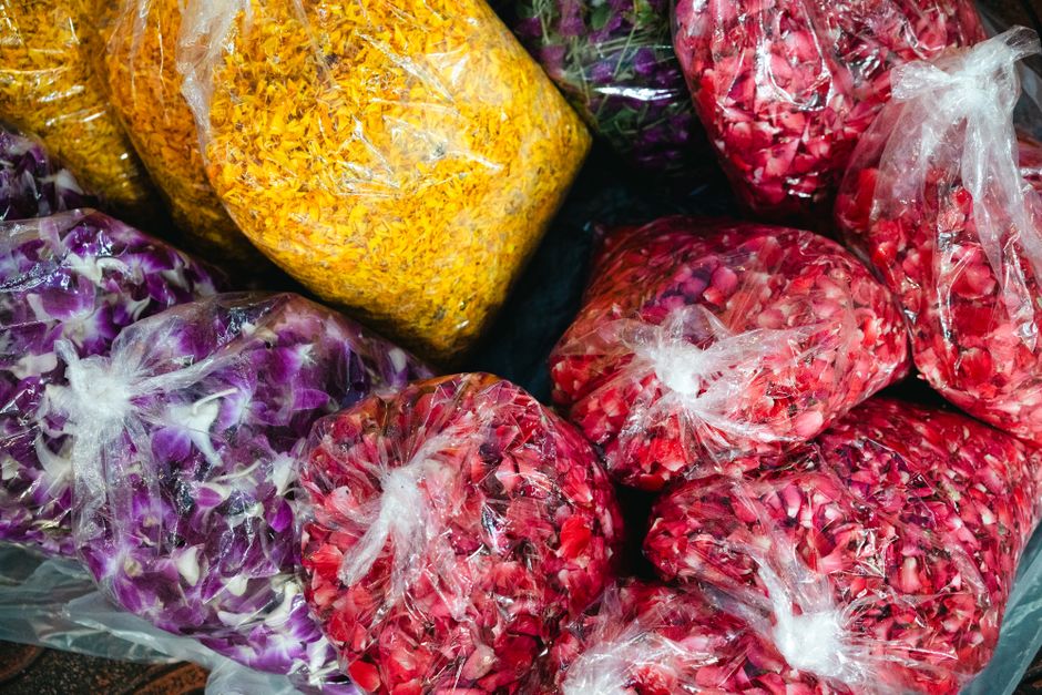 Close-up image of several transparent plastic bags filled with different types and colors of flower petals and dried flower materials. The bags contain vibrant yellow, purple, and red flower fragments