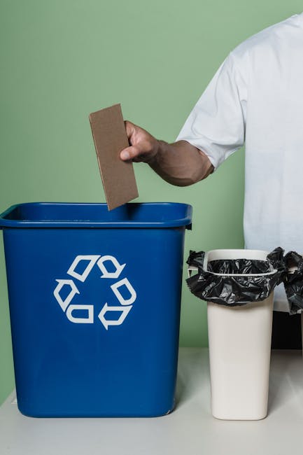 A person in a white shirt is holding a brown piece of cardboard over a blue recycling bin with a white recycling symbol, situated on a white surface against a light green wall. Next to the blue bin, t