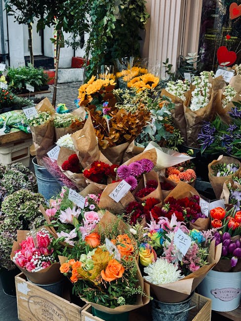 An outdoor display of a variety of fresh flowers arranged in multiple brown paper bouquets and metal buckets. Prominent flowers include bright yellow sunflowers, vibrant red and pink roses, purple and
