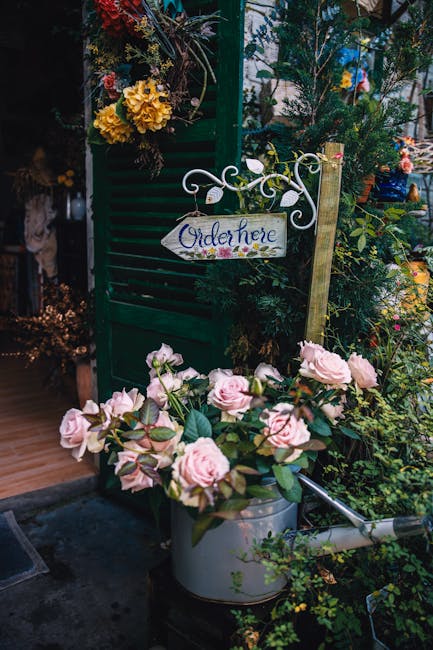 A charming outdoor display featuring a variety of fresh flowers, including pale pink roses with lush green leaves arranged in a silver metal watering can, positioned in front of a dark green wooden sh