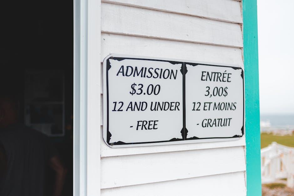 Close-up of a bilingual sign displaying admission prices, with the English side showing '$3.00 for 12 and under - free' and the French side reading '3,00$ 12 et moins - gratuit,' mounted on a white wo