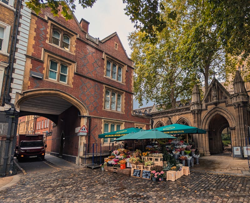 A street-level view of a flower stall set up under a green umbrella on a cobblestone pavement near a historic red-brick building with arched entryways and multiple windows. The stall displays an array