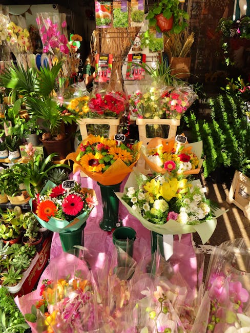 A vibrant display of fresh flower bouquets arranged on a pink cloth-covered table inside a florist shop. The bouquets include a mix of colorful blossoms such as bright yellow sunflowers, pink gerbera