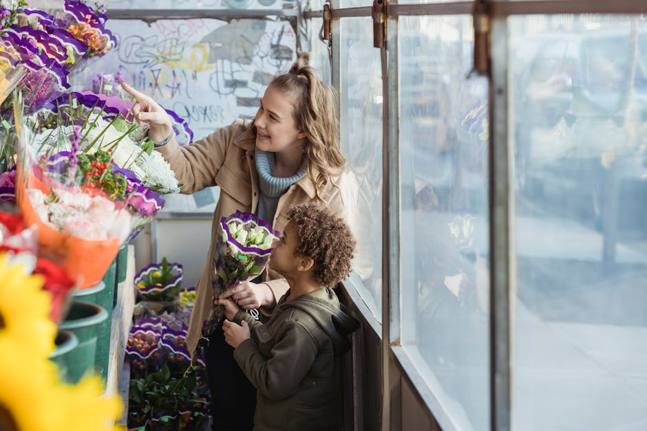 A woman and a young girl browsing fresh floral arrangements inside a greenhouse or flower shop, with the woman pointing at a vibrant display of purple and white flowers wrapped in decorative paper. Th