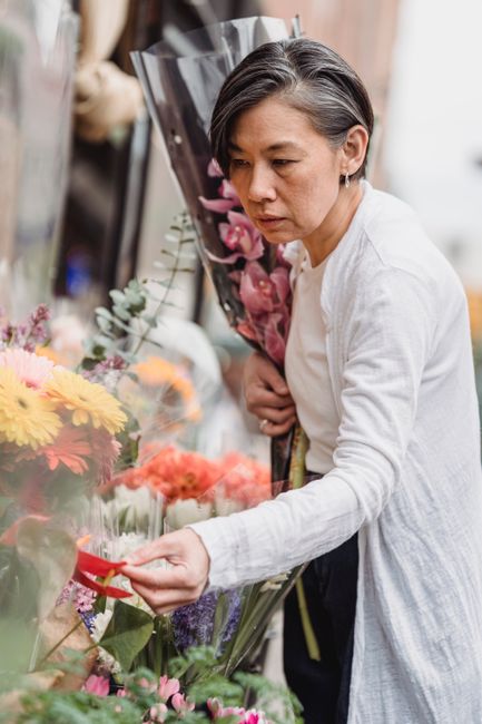 A woman with short, dark hair and a white long-sleeve top selecting a flower from a vibrant display of fresh, multicolored flowers at a florist shop. The floral arrangement includes bright yellow, pin