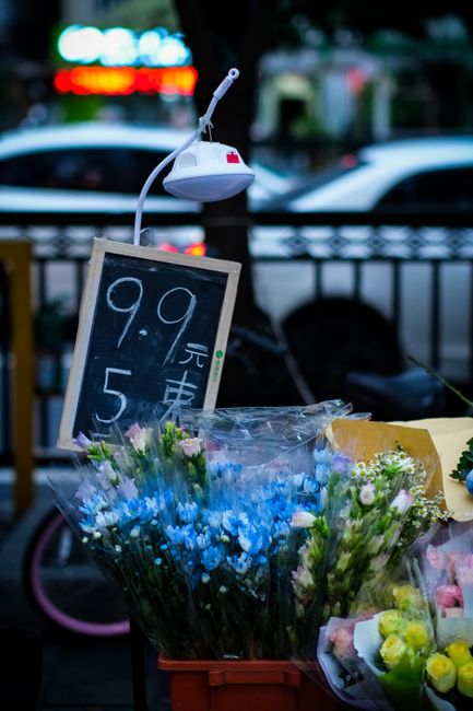 Close-up of a floral display outside a shop, featuring a variety of fresh flowers including blue and pink carnations, white daisies, and green foliage wrapped in transparent plastic. The flowers are a
