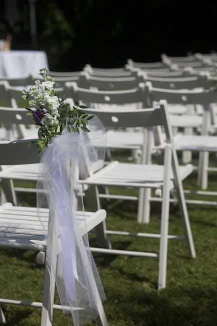 A delicate floral arrangement attached to a white wooden folding chair at an outdoor wedding venue, featuring a small bouquet of white lisianthus, purple lisianthus, and green foliage, wrapped in shee