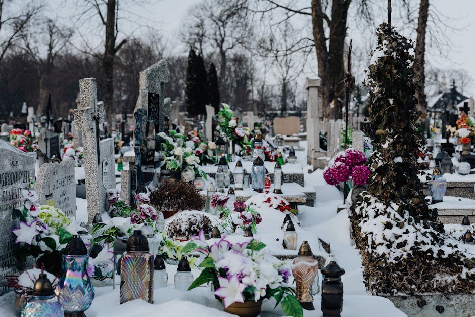 A snow-covered cemetery with numerous gravestones adorned with colorful floral arrangements, including pink and white hydrangeas, purple lilies, and mixed greenery, placed on and around the graves. La