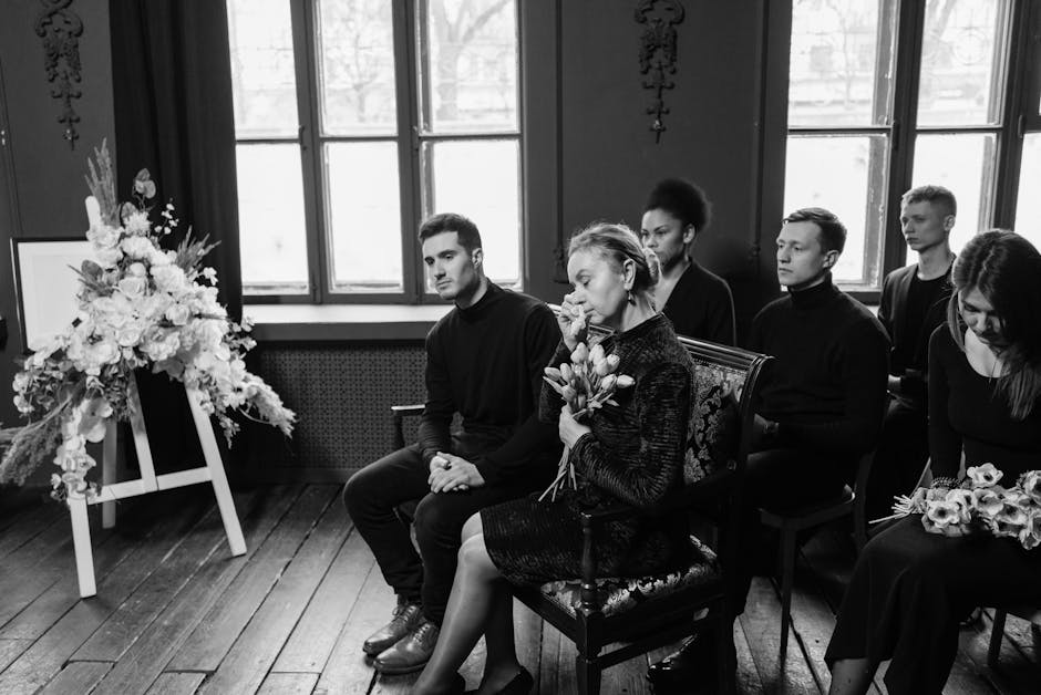 A black and white photo of six people seated indoors during a funeral or memorial service, with large windows in the background allowing natural light into the room. The central figure is a woman hold